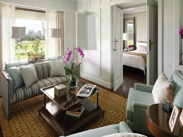 A living area with a striped sofa, armchairs and a wooden coffee table, with an open doorway to a bedroom and a window showing a green lawn and city buildings beyond.