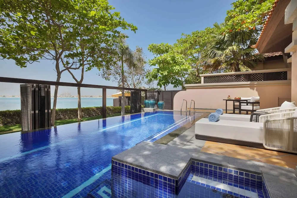 A long blue-tiled private pool sits beside white sun loungers on a terrace, with trees and the sea visible beyond the railing.