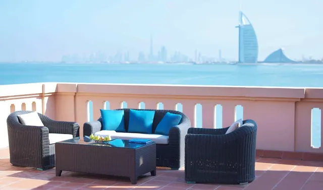 A terrace with wicker seating and blue cushions overlooks the sea, with the Burj Al Arab hotel and a city skyline in the distance.