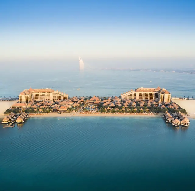 Aerial view of a resort with two large towers, low pavilion roofs, a sandy beach and overwater bungalows, with the Burj Al Arab visible in the hazy distance across the sea.