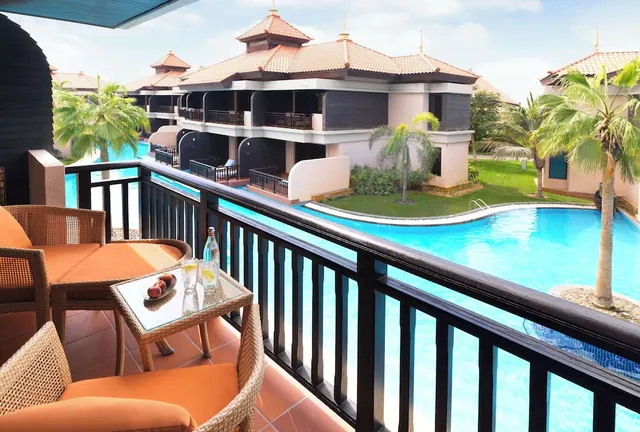 A balcony with wicker chairs and a glass side table overlooks a swimming pool, palm trees and low-rise resort buildings.
