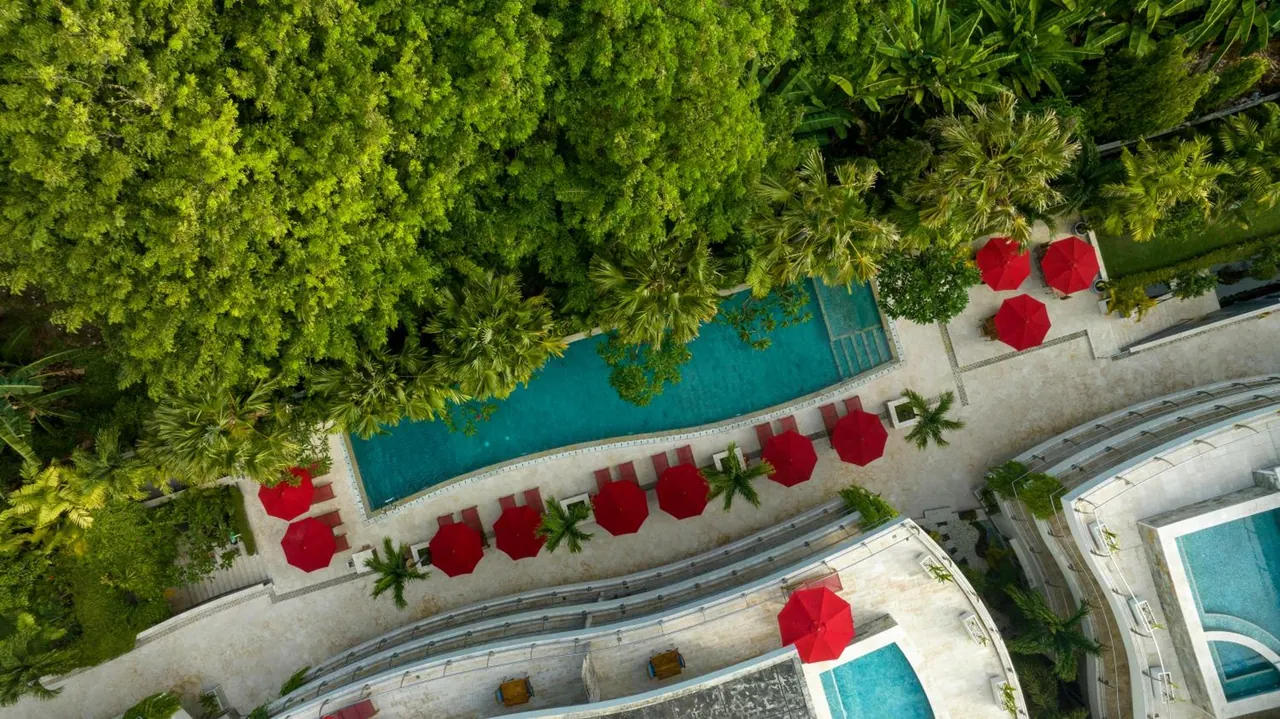 Aerial view of a pool with red umbrellas and sun loungers beside a dense tropical treeline, with other pool areas at the edges.