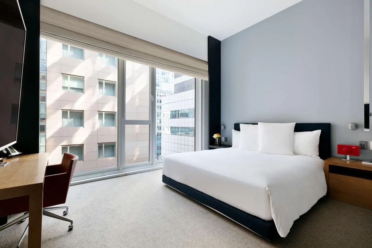 A bedroom with a made king bed, a wooden work desk with a red chair, a wall-mounted television, and large windows overlooking neighbouring buildings.