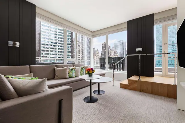 A living area with a grey sectional sofa, two round side tables and floor-to-ceiling windows overlooking high-rise buildings, with a raised wooden platform to the right.