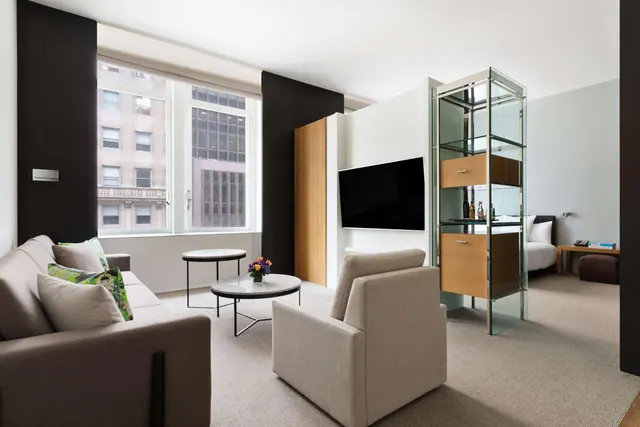 A living area with a grey sofa, an armchair, two round coffee tables, a wall-mounted television and a view of city buildings through the window, with a bed visible in the room beyond.