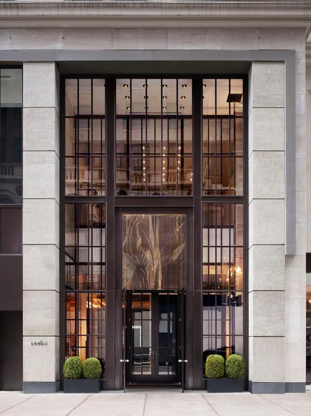A tall glass entrance with dark metal framing sits within a pale stone facade, with double doors and two clipped topiary shrubs in planters at street level.