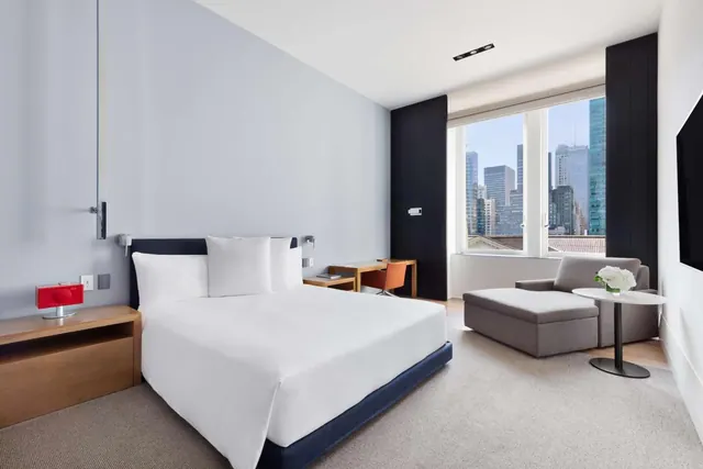 A bedroom with a king bed, a grey chaise and armchair, a wooden desk with an orange chair, and a window showing city skyscrapers.