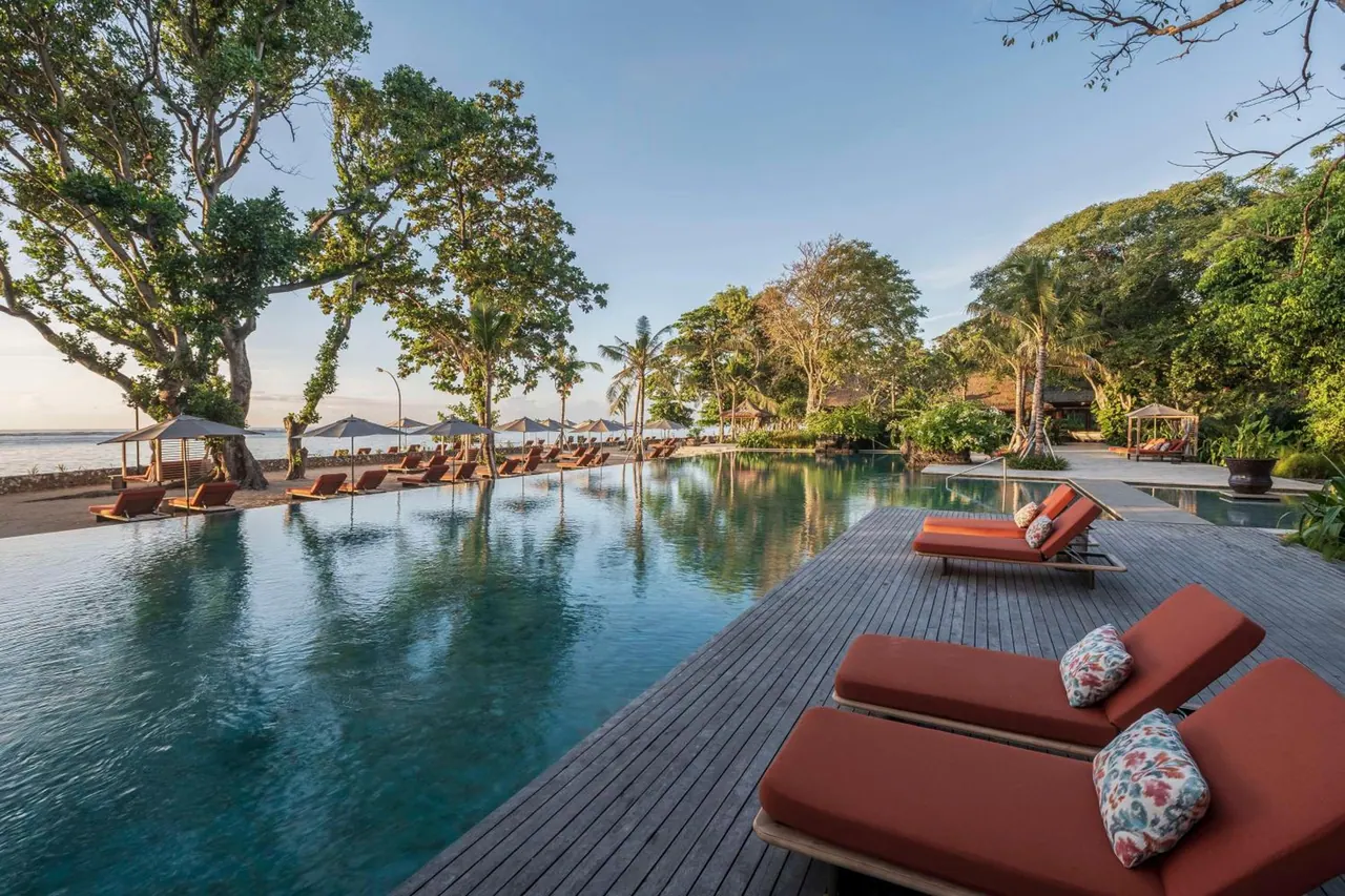 An infinity-edge pool with orange sun loungers and parasols runs along a sandy beach toward the sea, with tall trees on the right.