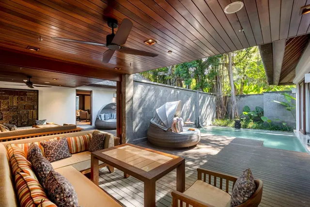 A covered outdoor terrace with a sofa seating area, a wooden coffee table, a wicker daybed and a small private plunge pool beside a grey wall with trees beyond.