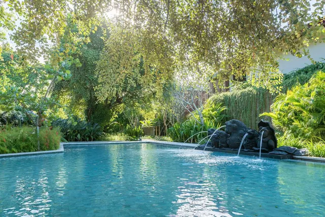 A pool with water spouting from dark stone sculptures at its edge, surrounded by tall trees and tropical plants in sunlight.