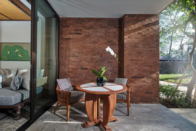A round wooden table with a white orchid and two wooden armchairs stands on a stone terrace beside a brick wall, with a sliding glass door to a sofa inside and a garden with trees beyond.