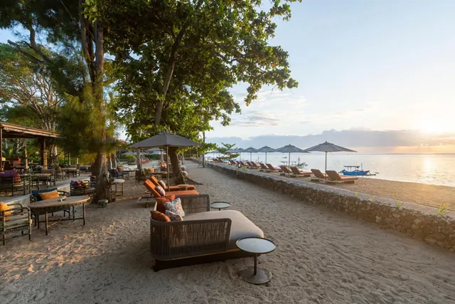 Sun loungers, shade umbrellas and an outdoor sofa sit on a sandy beach beside a stone wall, with the sea and a moored boat visible at sunset.