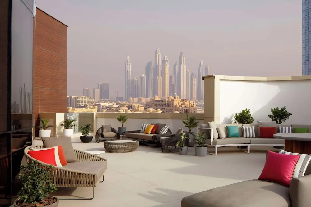 A rooftop terrace with outdoor sofas, cushions and potted plants overlooks a cluster of high-rise towers under a hazy sky.