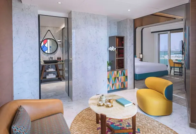 A living area with a tan leather sofa, yellow armchair and round marble coffee table, with an open doorway to a bedroom and bathroom and a view of water and buildings through the bedroom's glass doors.