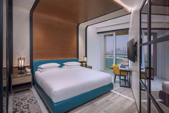 A bedroom with a made-up king bed, a work desk with a yellow chair, and a glass sliding door opening onto a balcony with a view of water and city towers.