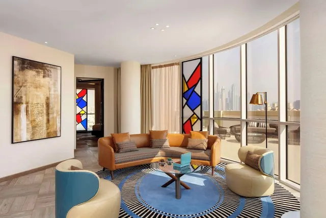 A living room with a tan leather sofa, a round blue and black patterned rug and curved floor-to-ceiling windows looking out onto a balcony and a city skyline.
