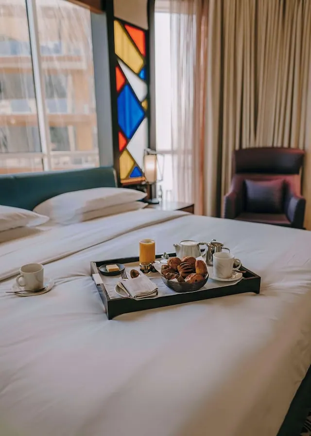 A breakfast tray with pastries, coffee cups and a teapot sits on a made bed beside a window and an armchair.