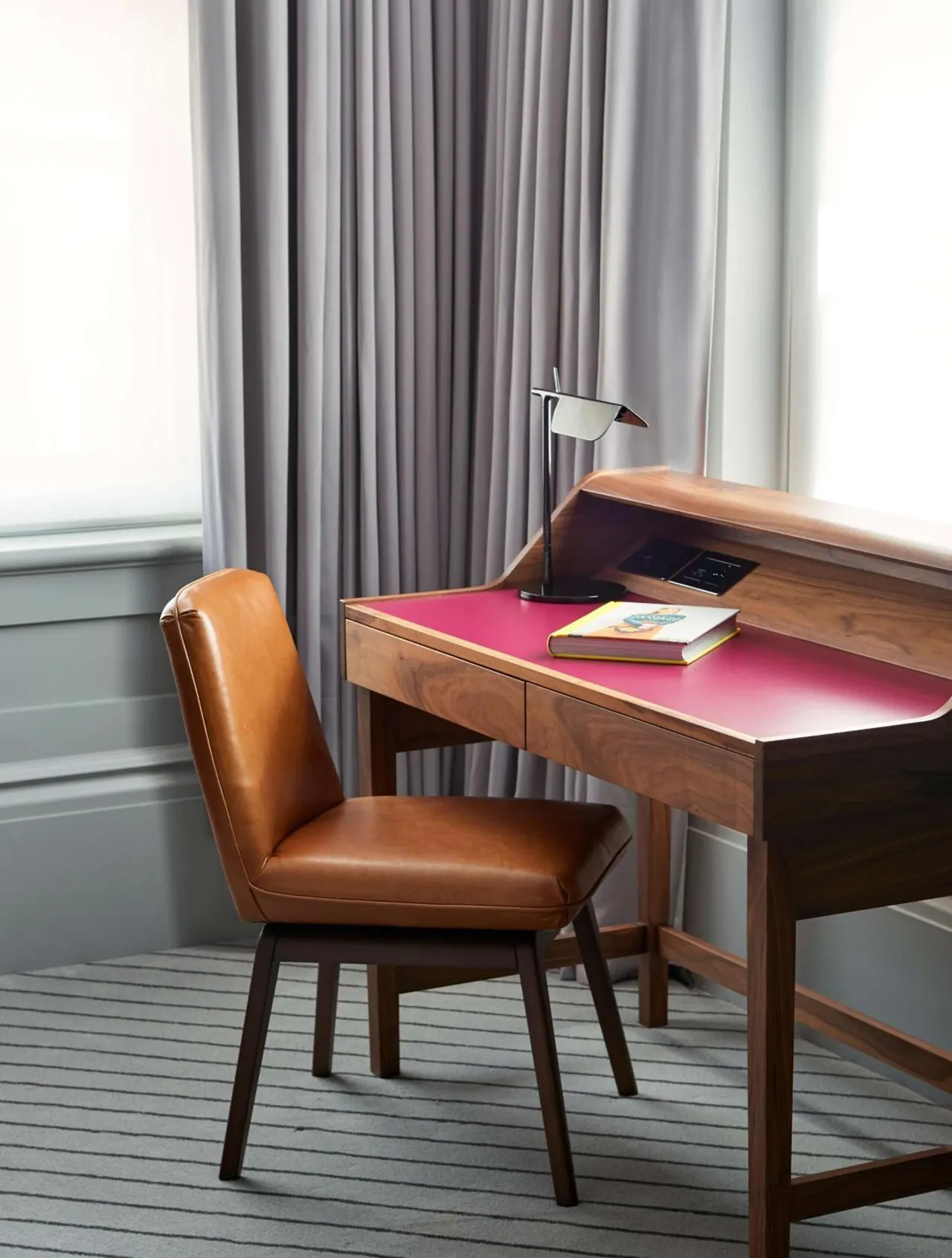 A walnut writing desk with a pink work surface, a black desk lamp, a book and a brown leather chair sit in front of grey curtains and a window.