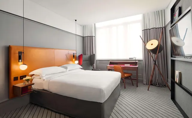A bedroom with a made king bed, orange leather headboard, a grey armchair, a writing desk with an orange chair, a floor lamp and a wall-mounted television.