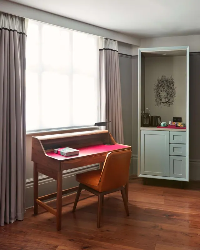 A wooden writing desk and orange leather chair stand by a window with grey curtains, with a cabinet holding a kettle and coffee machine on the right.