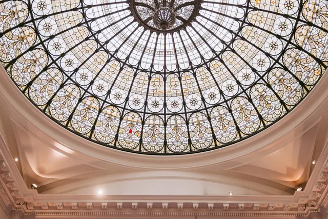 An ornate glass dome with a dark iron framework and a cream-coloured cornice sits above an interior space.