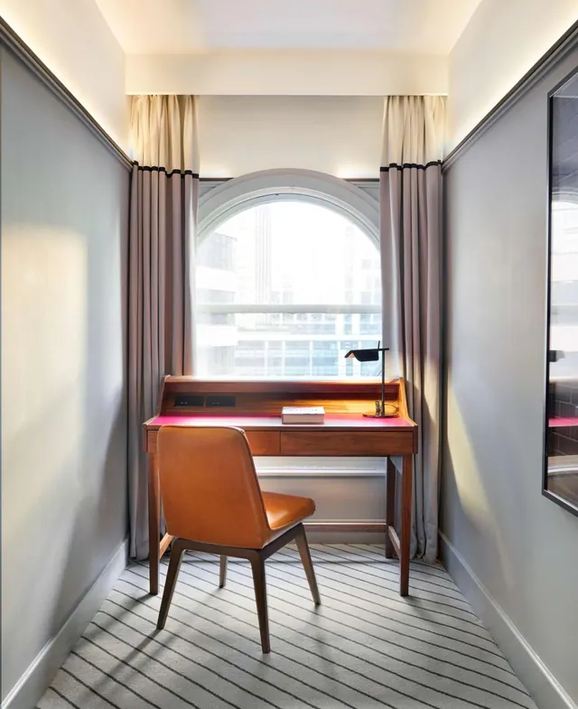 A wooden desk with a brown leather chair stands in front of an arched window with beige curtains, beside a striped grey carpet.