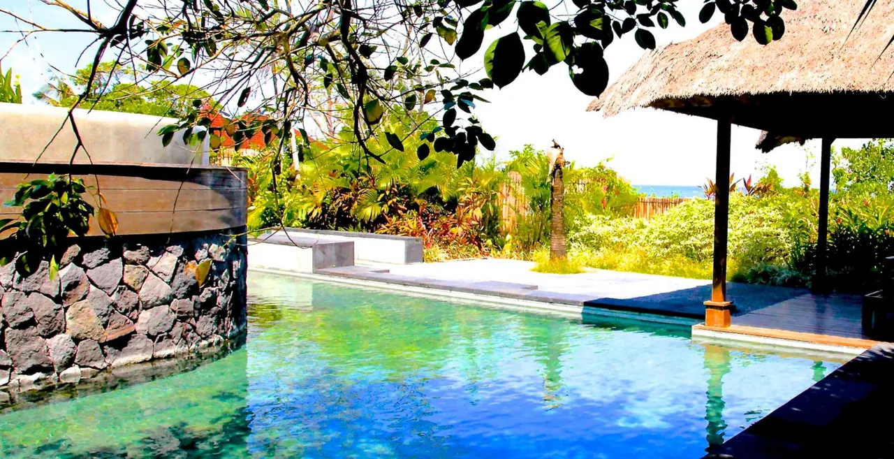 A pool with a stone wall on the left and a thatched pavilion on the right, with tropical garden and a strip of sea beyond.