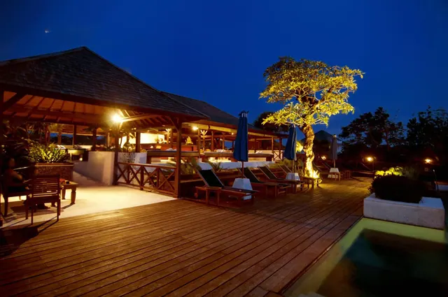 A timber deck with sun loungers and closed blue umbrellas runs beside a lit pavilion, an illuminated tree and a pool edge at night.