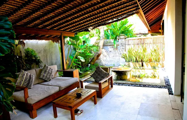 A covered outdoor seating area with a wooden sofa, armchairs and a low coffee table overlooks a garden with a stone water feature.