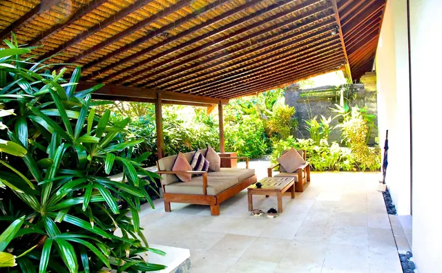 A covered terrace with a wooden daybed, cushions, an armchair and a low table, opening onto a tropical garden.