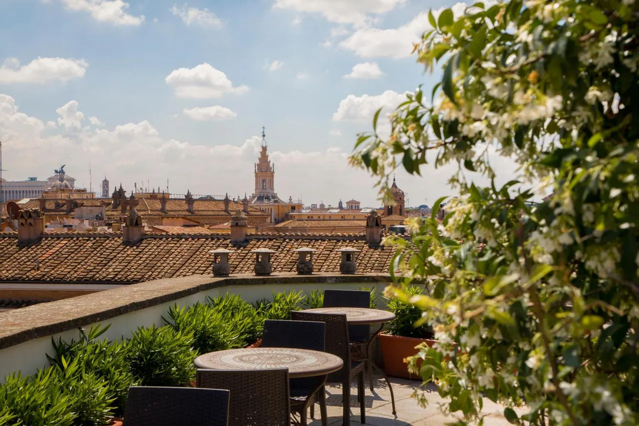 A rooftop terrace with round stone-topped tables and wicker chairs overlooks terracotta rooftops, a church bell tower and the Vittoriano under a partly cloudy sky.