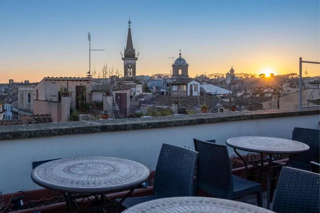 Mosaic-topped tables and wicker chairs sit on a rooftop terrace overlooking old Roman rooftops, church towers and a dome at sunset.