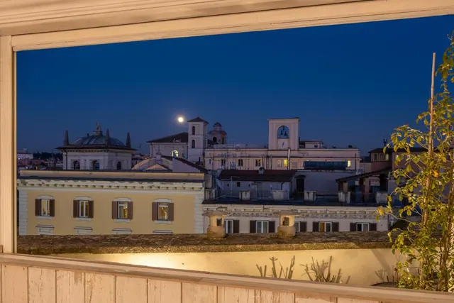 A night view over Rome rooftops, framed by a wooden balcony or window edge, showing a domed building, a church tower and a moon in the dark blue sky.