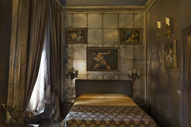 A bedroom with a made-up bed, three framed paintings on a textured mirror-tile wall, wall sconces, and heavy curtains beside a window.
