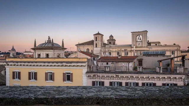 A dusk view over Rome's rooftops, with church domes and bell towers, seen from a stone terrace edge in the foreground.