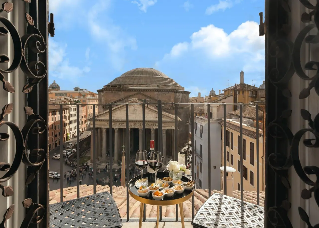 A balcony with wrought iron railings and a small table holding wine glasses and snacks overlooks the Pantheon dome and facade.