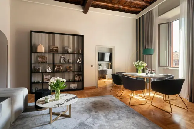 A living area with a bookshelf, a marble coffee table, a sofa and a dining table with chairs, with a window showing neighbouring buildings and a television visible through a doorway.