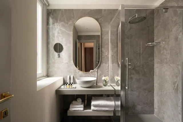 A bathroom with grey marble walls, an oval gold-framed mirror, a white basin on a wall-mounted shelf, folded towels below, and a glass-enclosed walk-in shower.