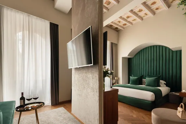 A double bed with a green velvet headboard sits beside a window with sheer curtains, under a painted coffered ceiling, with a wall-mounted television and a small table holding wine glasses.