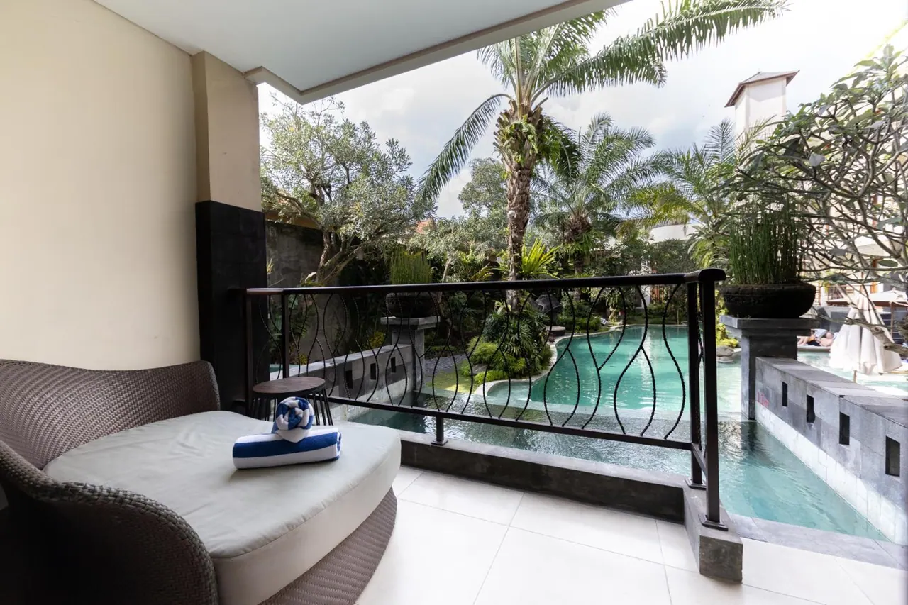 A balcony with a wicker daybed holding a folded towel, a metal railing, and a view over a curved swimming pool and palm trees.
