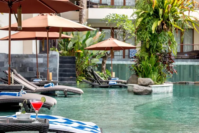 A turquoise hotel pool with brown parasol umbrellas and sun loungers, a cocktail on a table in the foreground, and tropical plants beside the water.
