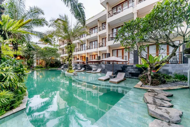 A large swimming pool with sun loungers and shade umbrellas sits in front of a multi-storey hotel building with palm and frangipani trees.