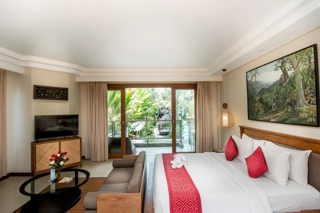 A bedroom with a king bed, wall-mounted television, sofa and glass coffee table, with sliding glass doors opening onto a balcony view of tropical garden.