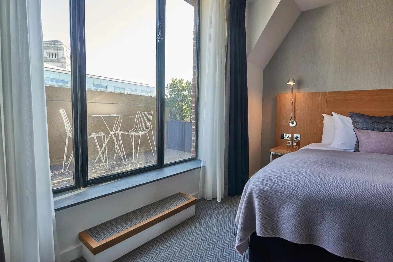 A double bed with a lavender throw sits beside a large window looking onto an outdoor terrace with a white table and chairs.