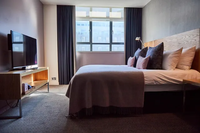 A double bed with cushions and a throw sits opposite a wall-mounted television, with a window looking onto a building across the street.