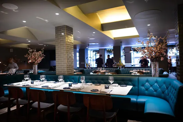 A dining room with teal tufted banquette seating, wooden chairs and set tables with wine glasses, with staff visible in the background near a bar.