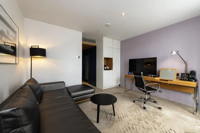 A hotel room living area with a black leather sofa, a floor lamp, a television on a wooden desk with an office chair, and a coffee machine beside white storage cabinets.