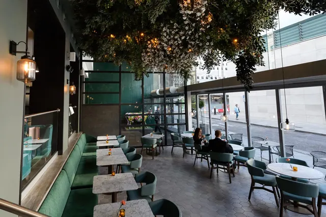 A restaurant dining area with green banquette seating, marble tables and hanging greenery, with two diners seated near floor-to-ceiling windows overlooking a street.