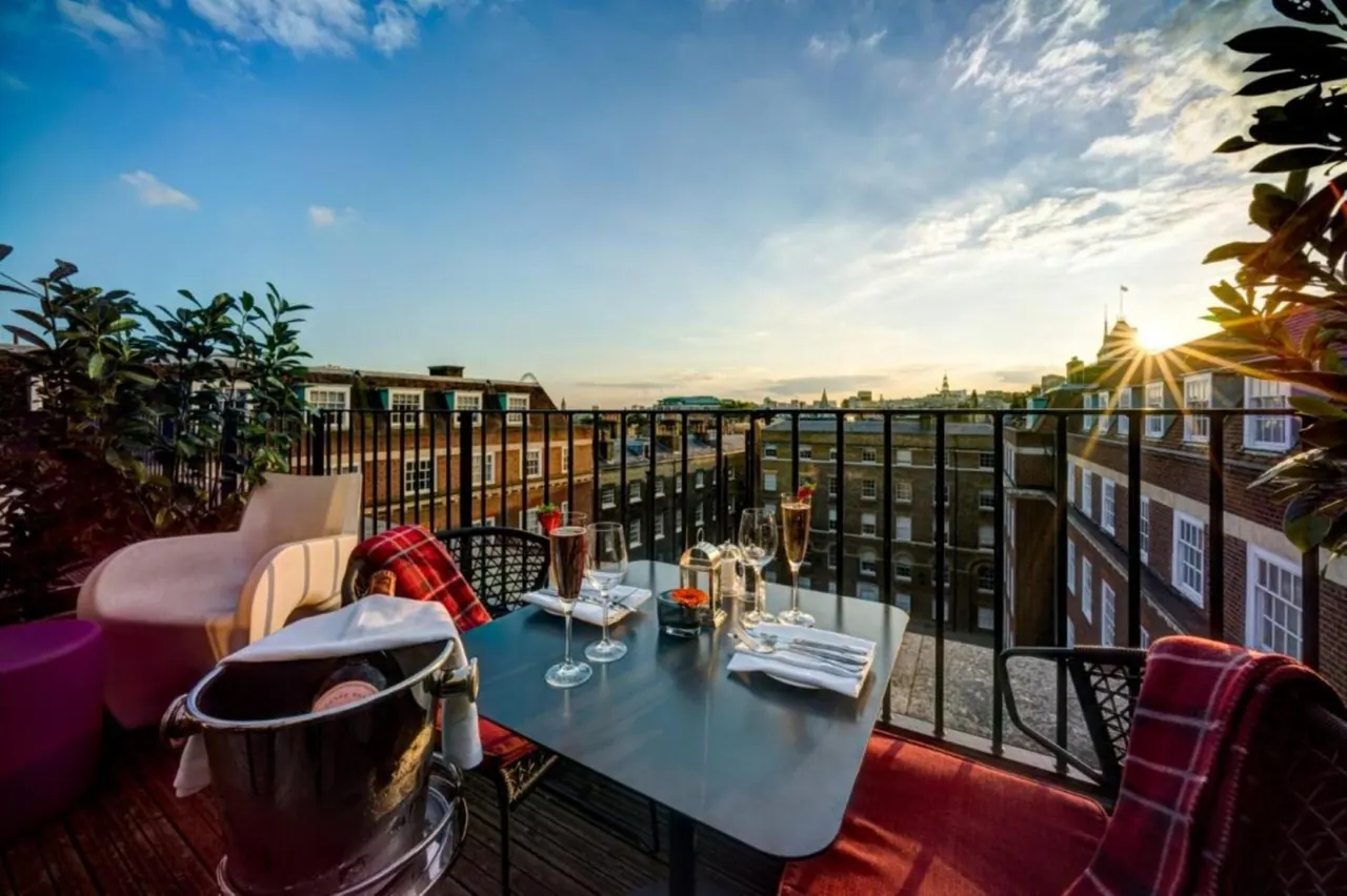 A set table with champagne glasses, a champagne bucket and folded napkins on a rooftop terrace overlooking brick buildings at sunset.