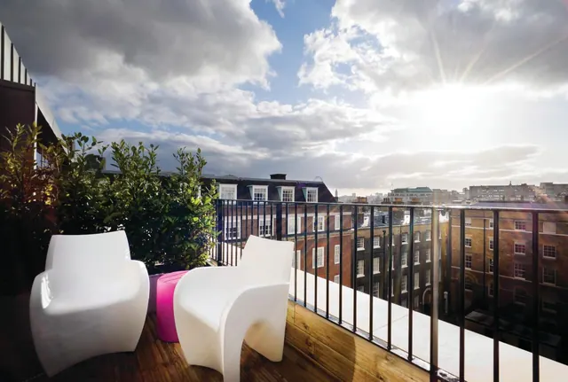 A rooftop terrace with two white plastic armchairs, a magenta stool, potted shrubs and a metal railing, overlooking brick buildings and a sunlit sky.